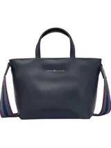 Cartera tipo tote de cuero sintético granulado color azul marino, con dos asas superiores y correa cruzada ajustable con cinta tejida a rayas azules y rojas. Presenta un cierre de cremallera principal y logo metálico de Tommy Hilfiger en el frente.