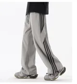 Pantalón deportivo gris claro, estilo cargo o jogging, con franjas negras verticales en los laterales de las piernas. Posee tobillos regulables y cintura ajustable.
