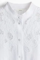 Blusa blanca de corte holgado en tejido liviano de lino y viscosa con canesú delantero en broderie. Cuello mao con frunces en la nuca, botones forrados adelante y mangas globo largas con puños finos abotonados. Bajo redondeado.
