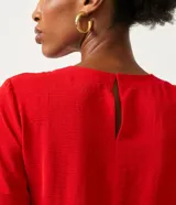 Blusa blanca de poliéster con cuello redondo, mangas cortas, cierre trasero en forma de gota, pequeñas aberturas laterales y acabados en pespuntes en las mangas y el cuello.