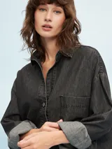 Camisa de denim negro con corte oversize, cuello clásico, cierre frontal con botones metálicos y bolsillo de parche en el pecho.