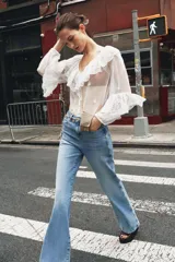 Blusa blanca semitransparente de escote pico y manga larga con puño elástico. Detalle de volantes y bordados a tono. Cierre frontal con botones.