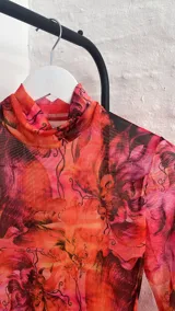 Vestido corto de microtul elastizado con estampado floral en tonos naranja, rosa y negro. Tiene cuello alto y mangas largas acampanadas.