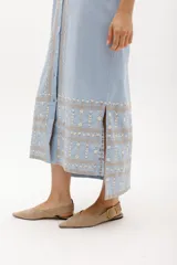 Vestido largo de lino en color celeste, con cuello camisero, manga corta y cierre frontal con botones. Presenta bordados decorativos en contraste en la pechera y en el ruedo.