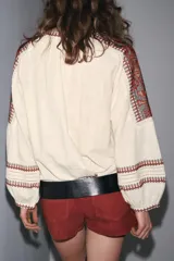 Blusa color crema de cuello redondo con escote pico y lazadas. Manga larga abullonada. Detalle de bordados combinados a contraste en cuello, mangas, pecho y puños. Cierre frontal con botones.