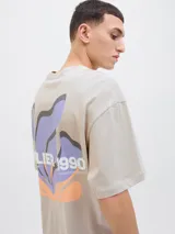 Remera beige de manga corta y cuello redondo con estampado en la espalda con la inscripción "Atelier © 1990" y un diseño abstracto en tonos lila y naranja.