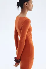 Vestido mini color naranja, entallado, de punto elástico con leve brillo, manga larga, escote en V, cuenta decorativa en el centro del busto y costura fruncida adelante para un efecto drapeado.