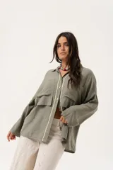 Camisa verde militar de manga larga con silueta oversize, dos bolsillos delanteros y corte levemente más largo en la espalda. Escote en V abotonado con detalle de galón picot a lo largo de la cartera y botones a tono. Isotipo bordado en la parte trasera.