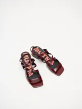 Sandalias de cuero charol color borgoña con tiras finas, taco cuadrado bajo y cierre con hebilla en el tobillo. Interior con estampado del logo de la marca en color naranja.