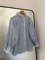Camisa holgada de rayas verticales celestes y blancas, con cuello alto y detalles de volados y puntilla blanca en el frente y puños.