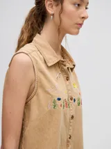 Camisa sin mangas de denim liviano color camel, con cuello camisero y botones frontales. Presenta bordados temáticos marinos en el pecho, incluyendo un sol, olas, una gaviota y vida marina.