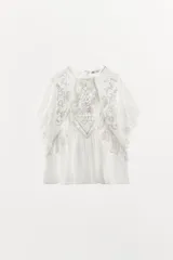 Top blanco de algodón con cuello redondo y manga corta con volados de encaje. Presenta bordados en el frente y cierre con botón en la espalda.