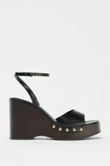 Sandalias negras con plataforma de madera, tacón de cuña y tira delantera con detalle de tachas doradas. Cierre mediante tira con pulsera al tobillo y acabado en punta cuadrada.