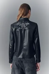 Camisa de símil cuero negra, de calce entallado, con cuello camisero y botones a presión con logo grabado. Presenta bordados florales con lentejuelas en los hombros y sobre los bolsillos frontales.