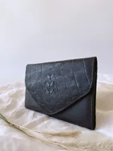 Billetera de cuero negro con textura tipo cocodrilo y cierre con broche. Tiene monedero externo y detalle de tres hojas grabadas en el frente.