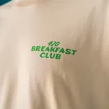 Remera oversize color beige con estampado en la espalda de una taza de café con cara sonriente, un cenicero con un cigarrillo y la frase "420 Breakfast Club. Wake and bake everyday".