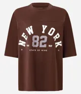 Remera de algodón color marrón oscuro con cuello redondo y mangas cortas, de corte oversized. Presenta un estampado frontal con el texto "NEW YORK 82" en tipografía universitaria y la frase "STATE OF MIND" en la parte inferior.