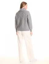 Buzo de punto grueso color beige, con cuello volcado y textura en relieve. Tiene mangas largas de corte hombro caído y puños anchos.