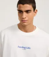 Remera de algodón blanca, corte oversized, con estampa gráfica en la espalda que incluye un teléfono antiguo, un CD, una cámara fotográfica, auriculares y un reproductor de CD portátil, junto al texto "Analog Life - NO EDITS -".