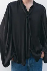 Camisa fluida negra de cuello redondo con lazada, manga larga tipo globo con pliegues en hombro y cierre frontal con botones ocultos por solapa.