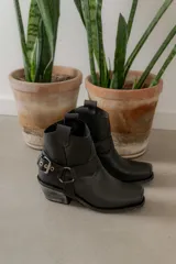 Botas cortas de cuero color marrón oscuro, con puntera cuadrada y caña baja. Presentan una correa con hebilla metálica alrededor del tobillo y un estribo desmontable.