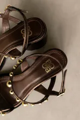 Sandalia de cuero color marrón con taco bloque medio y punta cuadrada. Presenta tachas esféricas doradas en las tiras y hebilla de ajuste en el tobillo.