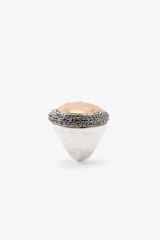Maxi anillo metálico plateado con forma ovalada y pieza incrustada labrada dorada a contraste. Detalle de aplicación de brillos negros alrededor de la pieza dorada.