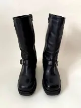 Bota de caña media de cuero vacuno color marrón oscuro, con punta cuadrada, cierre lateral y suela track negra. Presenta detalles de hebillas metálicas plateadas en la caña y en la parte inferior del empeine.