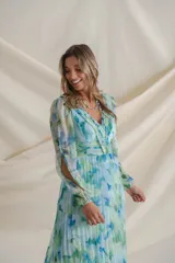 Vestido largo de gasa plisada con estampado floral en tonos celeste, verde y azul. Tiene escote cruzado con detalle de alforzas, mangas largas con abertura y falda plisada. Ideal para eventos de día.
