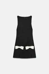 Vestido corto negro con cuello redondo y sin mangas. Presenta dos lazos blancos en la parte inferior delantera y aberturas frontales en el ruedo. Cierre trasero con cremallera metálica.