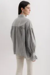 Camisa gris a rayas verticales blancas, de corte amplio, cuello alto con lazo y mangas largas abullonadas con puños elásticos.