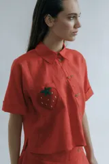 Camisa corta de manga corta color coral, con cuello clásico, doble cartera diagonal con botones de madera y botamangas marcados. Presenta un bolsillo con aplique de crochet hecho a mano con forma de frutilla.