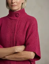 Capa o poncho tejido color verde esmeralda, con cuello polera alto que se cierra con un botón grande en el lateral. Presenta mangas cortas estilo japonesa y un calce amplio.