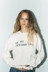 Sudadera corta color crudo de algodón, con cuello redondo, mangas largas y texto estampado en el frente que dice "IN MY YEEHAW ERA".