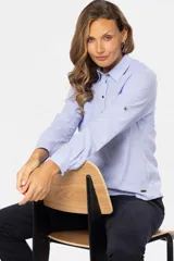 Camisa color beige de lino, de manga larga con botones metálicos personalizados y presilla en la manga. Tiene cuello clásico, cierre frontal parcial con botones y bolsillo de parche en el pecho.