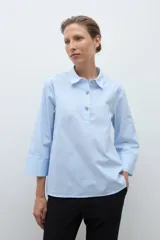 Camisa de popelina celeste con finas rayas blancas verticales, cuello camisero y abertura frontal con botones joya. Manga larga con puño ancho.