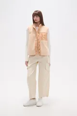 Chaleco holgado confeccionado en poliéster y corderito en tonos beige y crema. Presenta cuello alto, cierre frontal con botones a presión y un bolsillo con cierre en el pecho. Combina texturas de corderito con paneles de tela lisa.