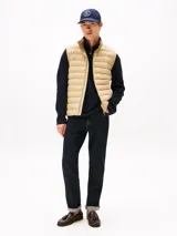 Chaleco acolchado color beige, con cuello chimenea y cierre frontal de cremallera. Presenta costuras horizontales y logo de Tommy Hilfiger bordado en el pecho.