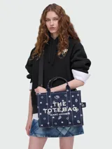 Bolso tipo tote de lona color azul marino con diseño gráfico estampado en color blanco. Incluye el nombre de la marca 'Marc Jacobs' y el modelo 'The Tote Bag' en el frente, con detalles de estrellas, corazones y flores. Posee doble asa superior, correa de hombro ajustable y costuras en contraste.