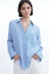 Camisa celeste y blanca a rayas verticales, de lino liviano, con cuello clásico, botones frontales, canesú de doble capa en la espalda, hombros caídos, mangas largas con puños abotonados, bolsillo de parche en el pecho y bajo redondeado.
