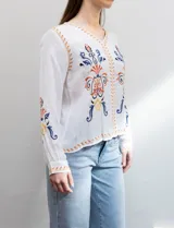 Blusa blanca de rayón con escote en V y mangas largas. Presenta bordados florales en tonos naranja, azul y negro en la parte delantera y en las mangas.