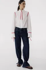 Camisa blanca de corte cropped con cuello mao y mangas largas abullonadas con lazos rojos en los puños. Presenta detalles de ribetes rojos en los hombros y lazos rojos en el cuello.