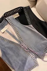 Chaleco negro de cuero con escote en V y cierre frontal con cordones. Presenta ojalillos metálicos plateados a lo largo de los bordes.