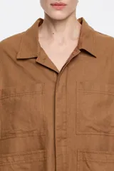 Chaqueta corta color marrón de corte recto, confeccionada en mezcla de lino y algodón. Tiene cuello camisero, botonadura frontal oculta, manga larga con pliegues en los codos, bolsillos plastrón en delantero y pecho, y pespunte a contraste en bolsillos, bajo y puños.