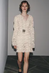 Vestido corto color crudo confeccionado en tejido tipo crochet con diseño geométrico floral. Tiene cuello solapa, abertura con botón en la parte superior y mangas largas acampanadas.