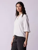 Blusa blanca de gasa marca Sioni, con cuello a la base y escote en V. Tiene abertura central con botones y mangas 3/4 con terminación fruncida.