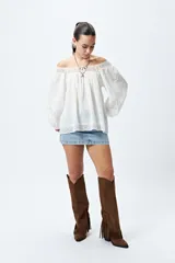 Blusa blanca de algodón con escote off-shoulder, mangas largas con puños elásticos y detalles de bordado y puntillas en escote y mangas.