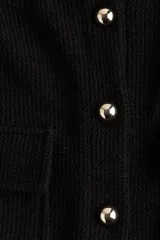 Top corto negro en tejido de punto waffle, con cuello redondo, manga corta, cierre frontal con botones dorados y bolsillos de parche con solapa y botón en el pecho.