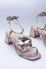 Sandalias de cuero color champagne, con taco cuadrado, tira delgada sobre los dedos y tobilleras para atar. Presentan un aplique de flor de cuero en la capellada.