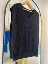 Chaleco negro de hilo con escote en pico, punto elástico en cuello, sisa y bajo. Parte trasera más larga que la delantera. Diseño en la parte delantera.